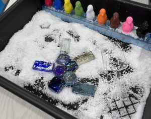 Small sensory tray filled with artificial snow, colorful glass gems, a clear scoop, and a row of multicolored penguin counters lined up on a number strip.