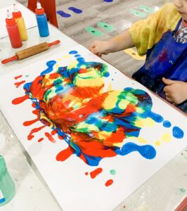 A child creates mitten symmetry art by folding paint on a large paper mitten shape, spreading bright red, yellow, blue, and green paint into a symmetrical pattern.