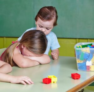 Preschool behavior moment showing one child taking a break at a table while another child stands nearby, illustrating early self regulation and peer awareness.